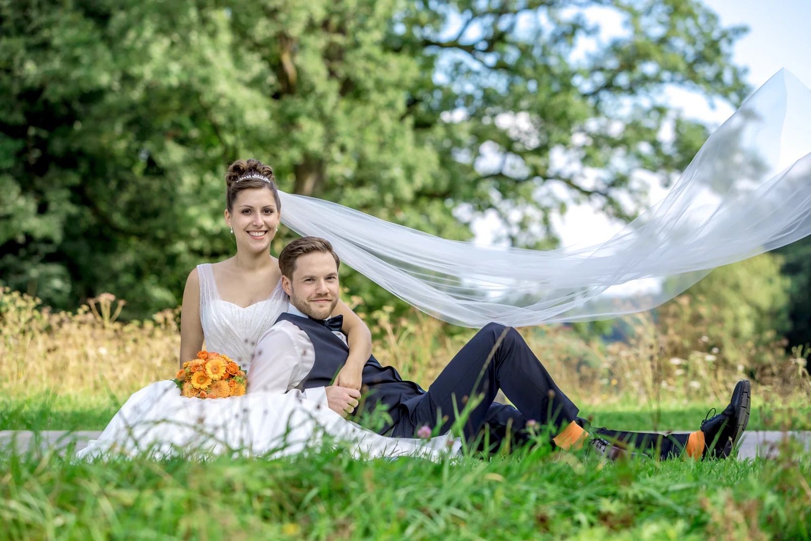 Das Brautpaar entspannt im Grünen mit wehendem Schleier – Hochzeitsreportage von Veronika und Andreas
