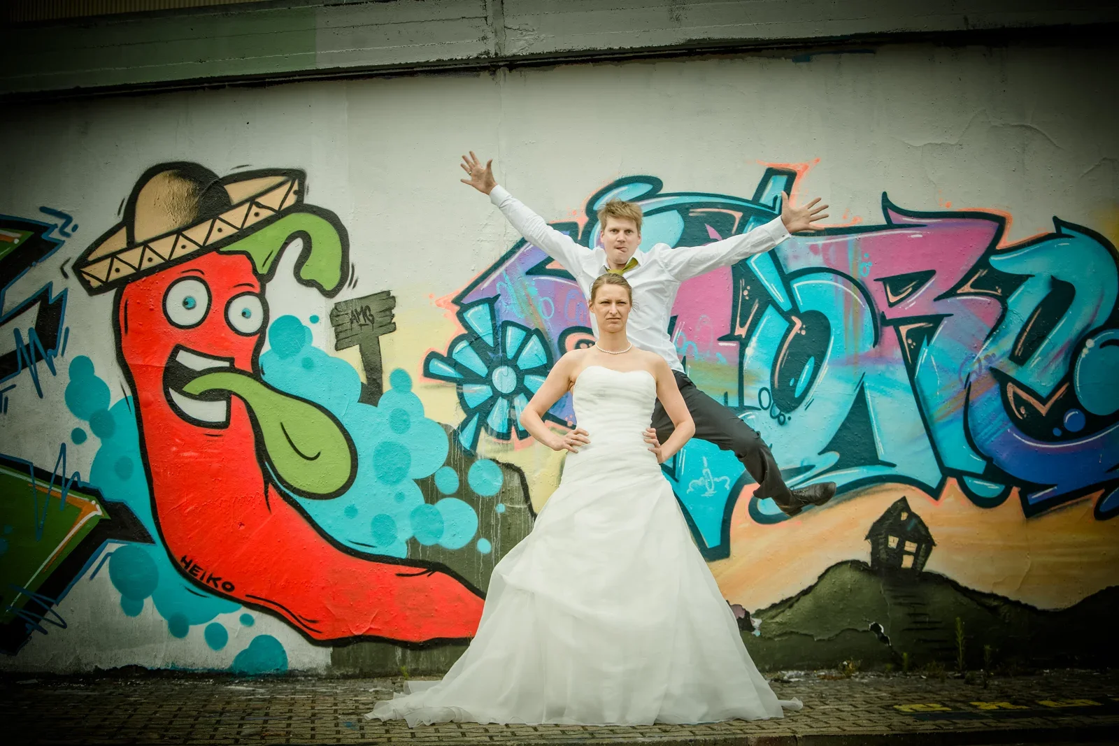 Viktoria im Vordergrund, Sebastian springt mit ausgebreiteten Armen – Hochzeit von Viktoria und Sebastian